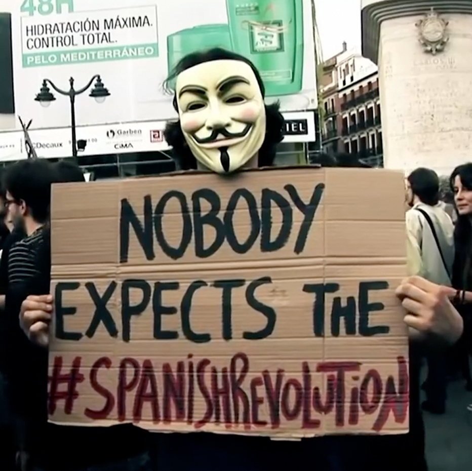 Person on street in Guy Fawkes mask holding a sign that says Nobody Expects The #SpanishRevolution. 